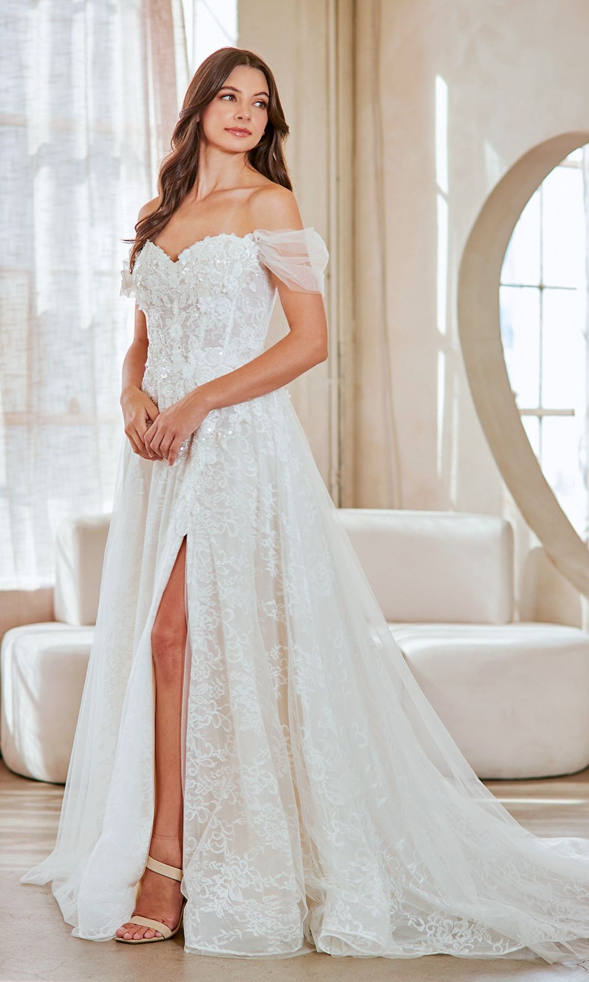 A woman in the Amelia Couture 2139 long wedding dress, featuring off-the-shoulder lace and a thigh-high slit, stands indoors near a large round mirror and cream sofas.